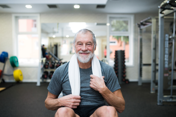 Fit senior man in sports clothing in gym resting after working out, towel around his neck