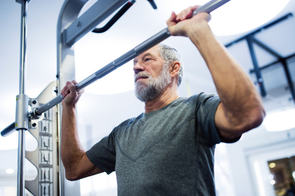 Fit senior man in sports clothing in gym working out, doing pull-ups on horizontal bar.