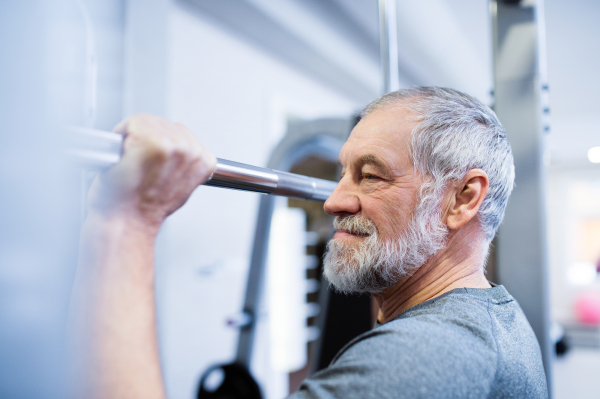 Fit senior man in sports clothing in gym working out, doing pull-ups on horizontal bar.