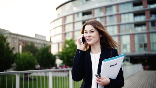 A happy young businesswoman with smartphone walking on the bridge in a city, holding files and making a phone call. Slow motion.