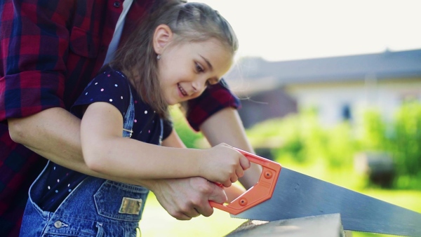 A close up of small girl and her unrecognizable father with a saw outside, cutting wood. Slow motion.