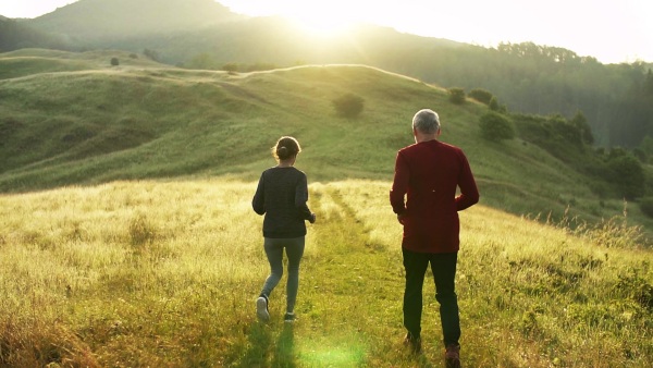 Rear view of senior sporty couple running outdoors on meadow in nature at sunrise. Slow motion