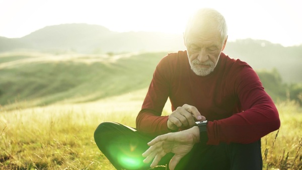 Senior man runner sitting on meadow outdoors at sunrise in nature, using smartwatch. Slow motion.