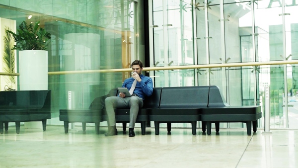 A young businessman with tablet and coffee sitting on a bench in a modern building, reading. Slow motion.