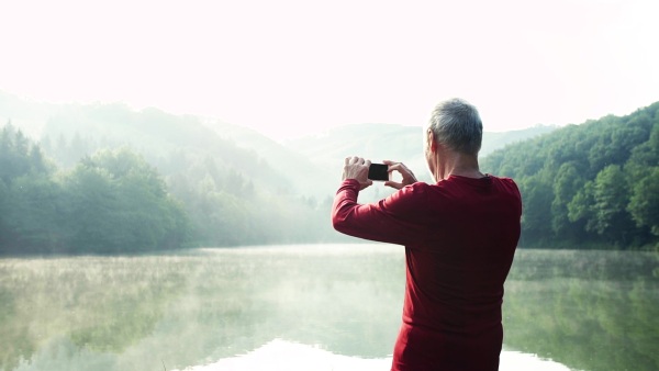 Senior man runner standing by the lake outdoor in foggy morning in nature, taking photograph. Copy space. Slow motion. Rear view.