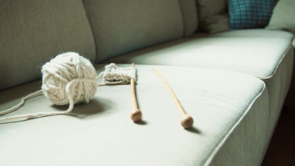 A ball of white yarn, wooden needles and a piece of knitted garment on the sofa at home. Close up.