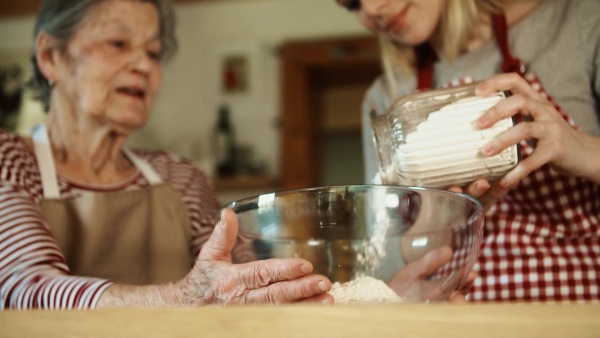 A happy elderly grandmother with an unrecognizable adult granddaughter putting flour in a bowl at home. Slow motion.