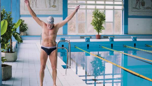 Senior man standing by the indoor swimming pool, stretching. Active pensioner enjoying sport.