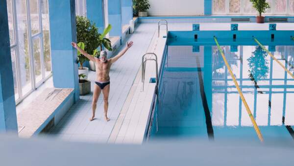 Senior man standing by the indoor swimming pool, stretching. Active pensioner enjoying sport.