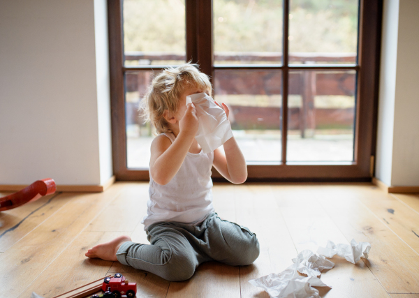 Small sick boy with cold at home sitting on floor, sneezing and blowing nose.