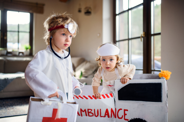 Two small children with doctor uniforms and toy ambulance car indoors at home, playing.