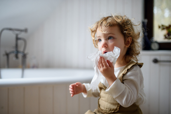 Small sick girl with cold at home sitting on ground, blowing nose. Copy space.