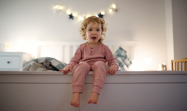 Small toddler girl sitting on edge of bed indoors at home, looking at camera.