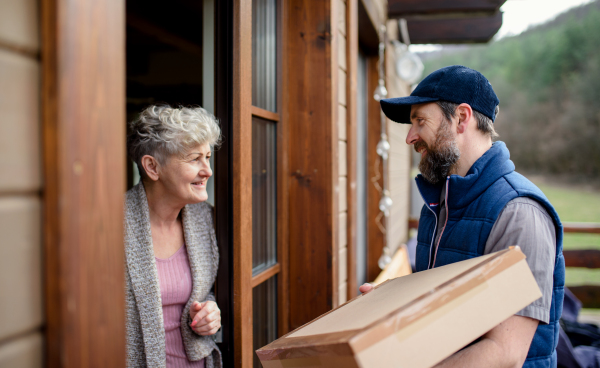 Man courier delivering parcel box to senior woman, standing by doorstep.