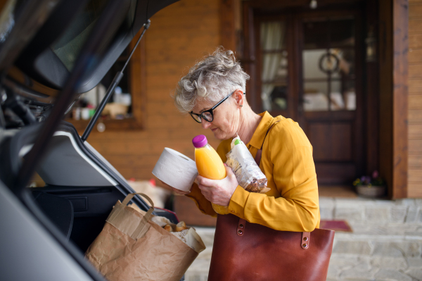 Senior woman with shopping in paper bag standing outdoors by car.