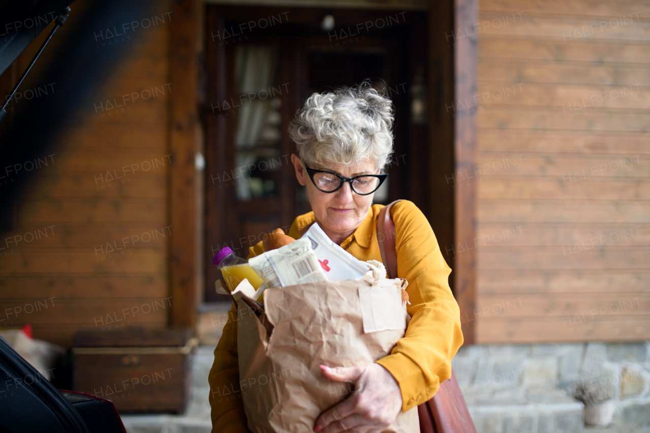 Senior woman with shopping in paper bag standing outdoors by car.