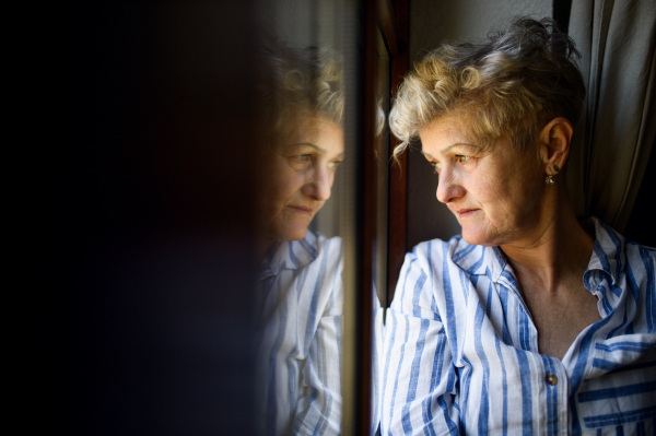 Sad senior woman standing indoors by window at home, corona virus and quarantine concept. Copy space.