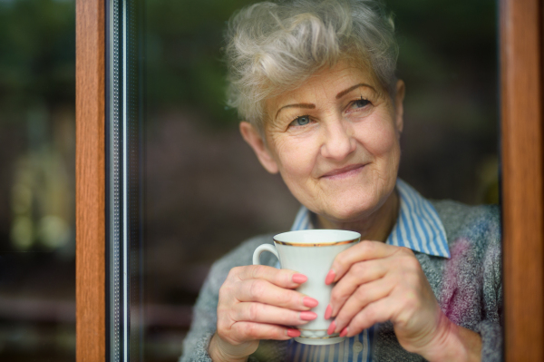 Sad senior woman with coffee looking out indoors at home, corona virus and quarantine concept.