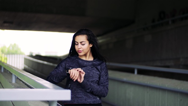 Beautiful young woman standing under the bridge in the city, using smartwatch.