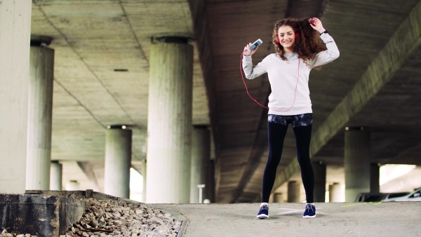 Beautiful young woman with smartphone and headphones under the bridge in the city, listening to music and dancing.