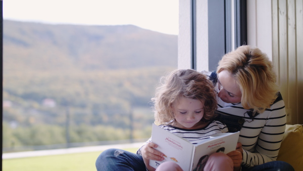 A cute small girl with mother sitting by window indoors at home, reading book.