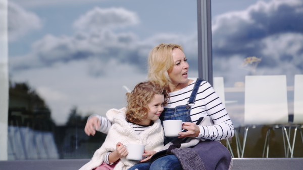 A cute small girl with mother sitting by window on patio outdoors, talking.