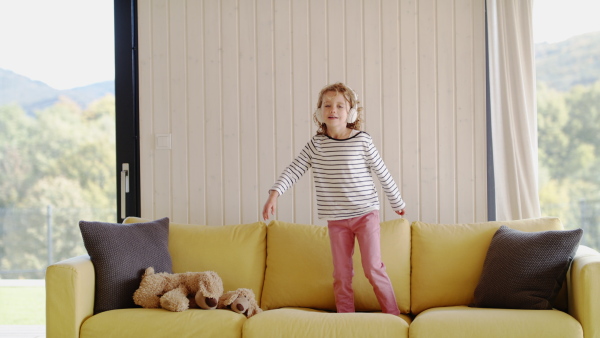 A cheerful cute small girl with headphones on sofa indoors at home, jumping.