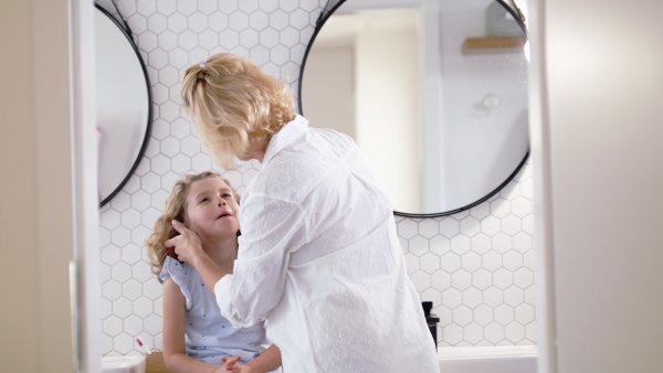 A cute small girl with unrecognizable mother in bathroom indoors at home, combing hair.