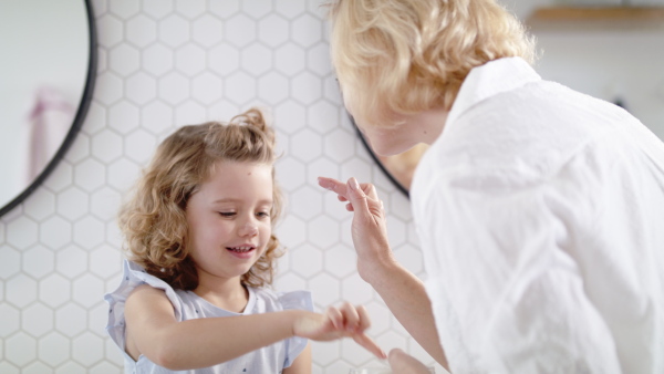 A cute small girl with mother in bathroom indoors at home, putting cream on face.