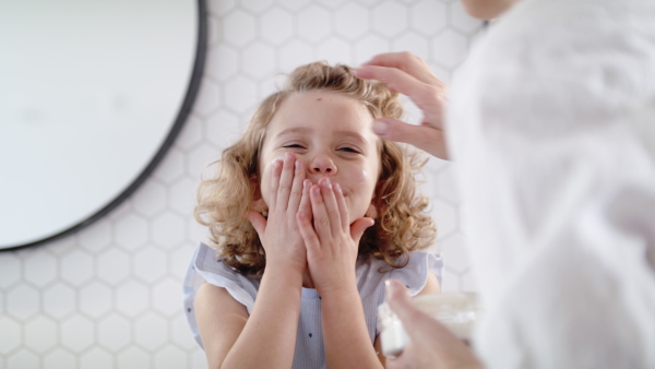 A cute small girl with mother in bathroom indoors at home, putting cream on face.