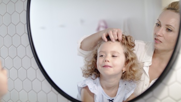 A happy cute small girl with mother in bathroom indoors at home, looking in mirror.