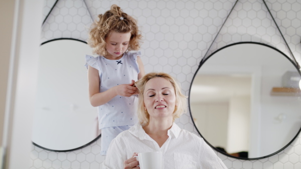 A cute small girl with mother in bathroom indoors at home, braiding hair.