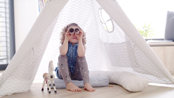 A cute small girl sitting in bedroom indoors at home, playing with binoculars.