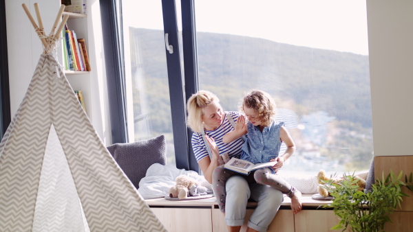 A cute small girl with mother sitting by window indoors at home, reading book.