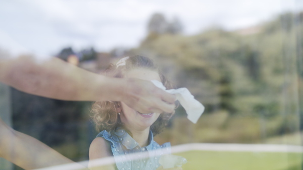 A cute small girl helping with housework indoors at home, cleaning window. Shot through glass.