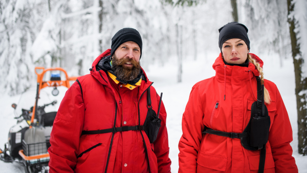 Portrait of woman and man paramedic from mountain rescue service outdoors in winter in forest, looking at camera.