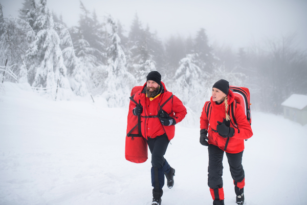 Man and woman paramedics from mountain rescue service running outdoors in winter in forest.