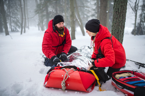 Paramedics from mountain rescue service provide operation outdoors in winter in forest, injured person in stretcher.