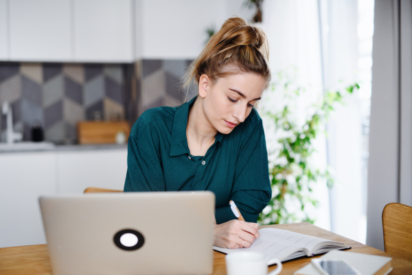 A young woman student with laptop writing notes at home, home office and learning.