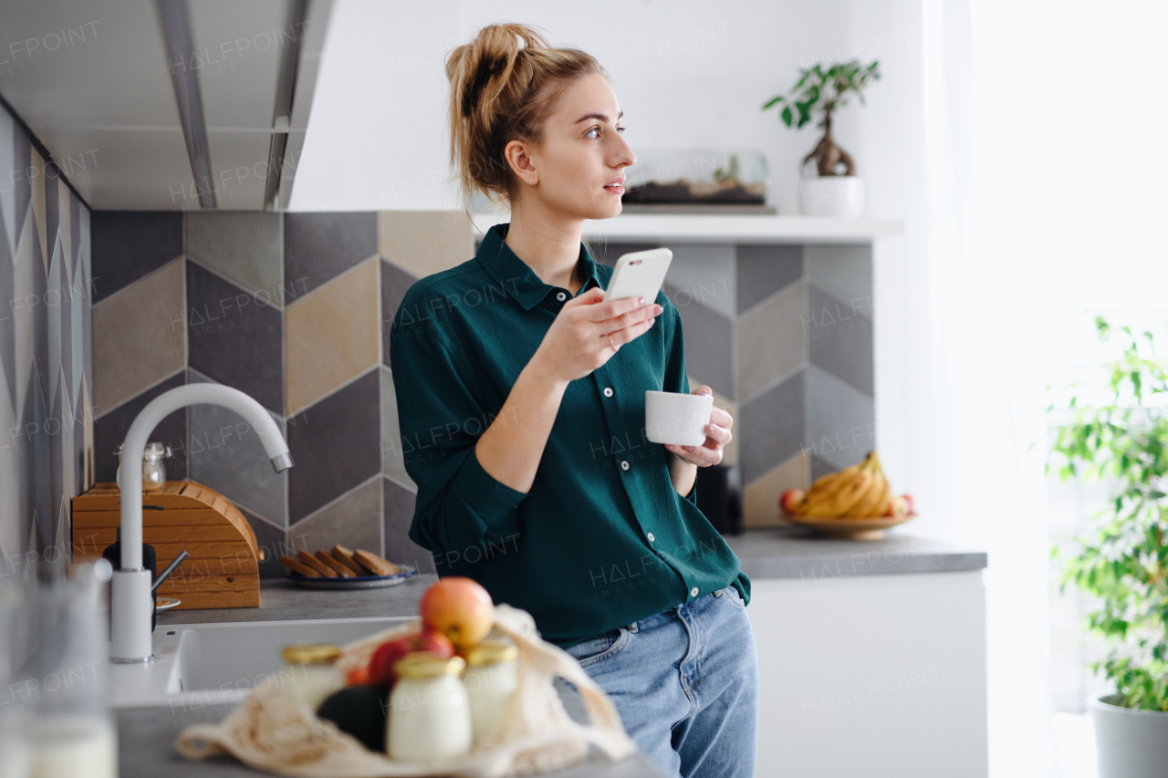 A portrait of young woman student with coffee at home, using smartphone.