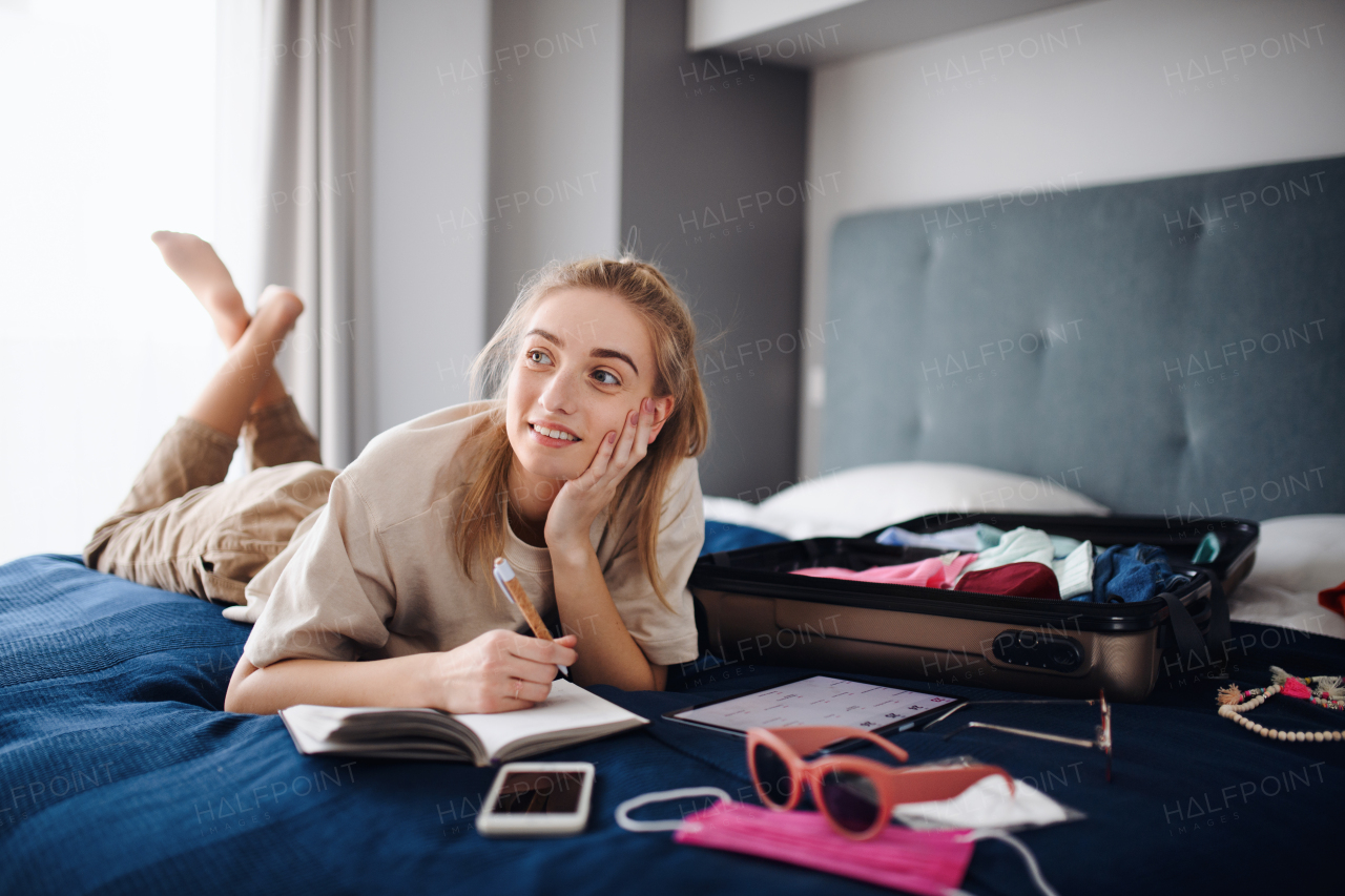 A portrait of young woman writing notes and packing clothes into suitcase indoors at home, planning holiday.