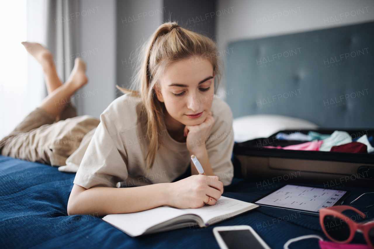 A portrait of young woman writing notes and packing clothes into suitcase indoors at home, planning holiday.