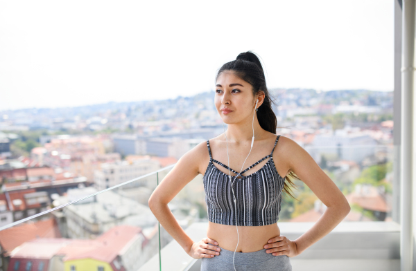 A portrait of young sport woman with earphones standing on balcony outdoors in city, resting.