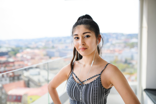 A portrait of young woman with earphones standing on balcony outdoors in city, looking at camera.