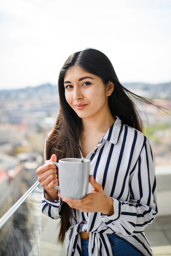 A portrait of young woman with coffee standing on balcony outdoors in city, looking at camera.