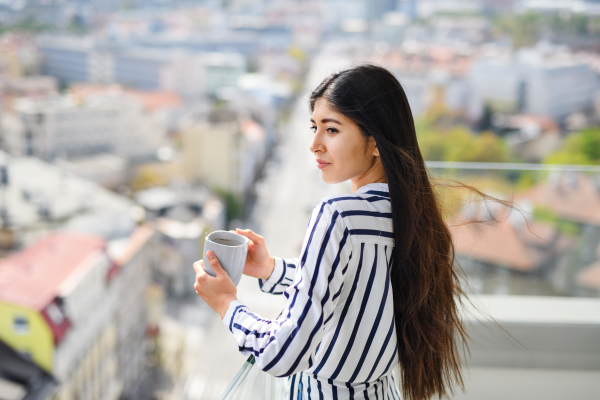 A portrait of young woman with coffee standing on balcony outdoors in city, looking at cityscape.