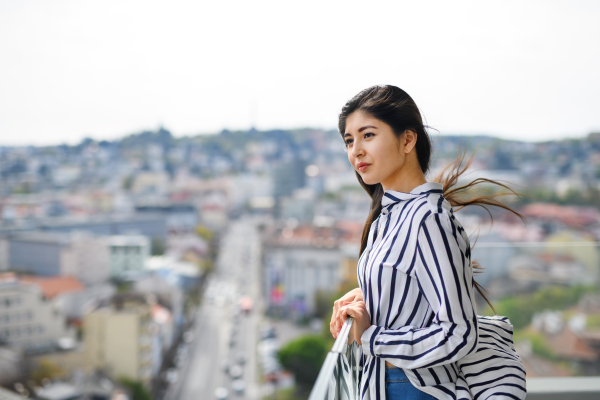 A portrait of young woman standing on balcony outdoors in city, looking at cityscape.