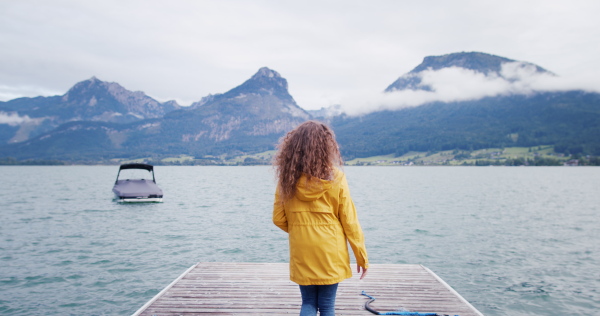 Rear view of young woman tourist in yellow jacket walking on a pier by lake.