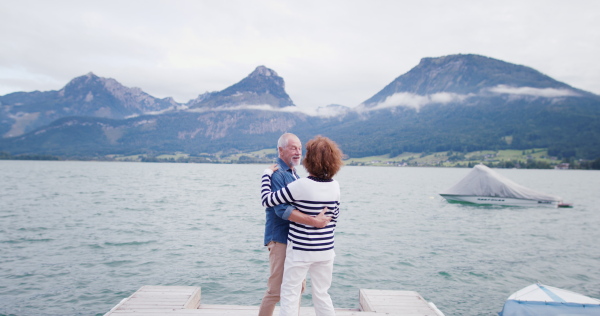 Rear view of senior pensioner couple tourists dancing on pier by lake, holding hands.