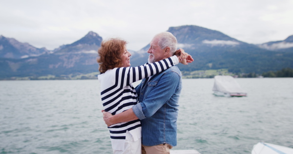 Side view of senior pensioner couple tourists hugging on pier by lake, holding hands.